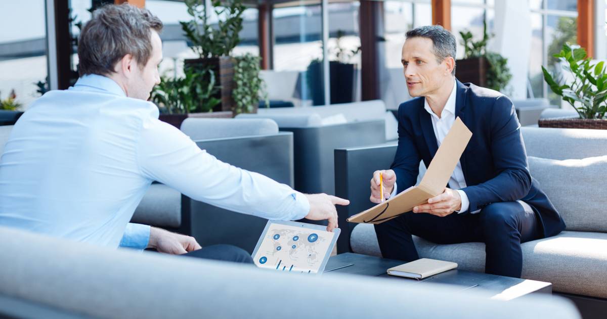 Man in pak wijst naar document in zijn hand aan man tegenover hem met een laptop voor zich